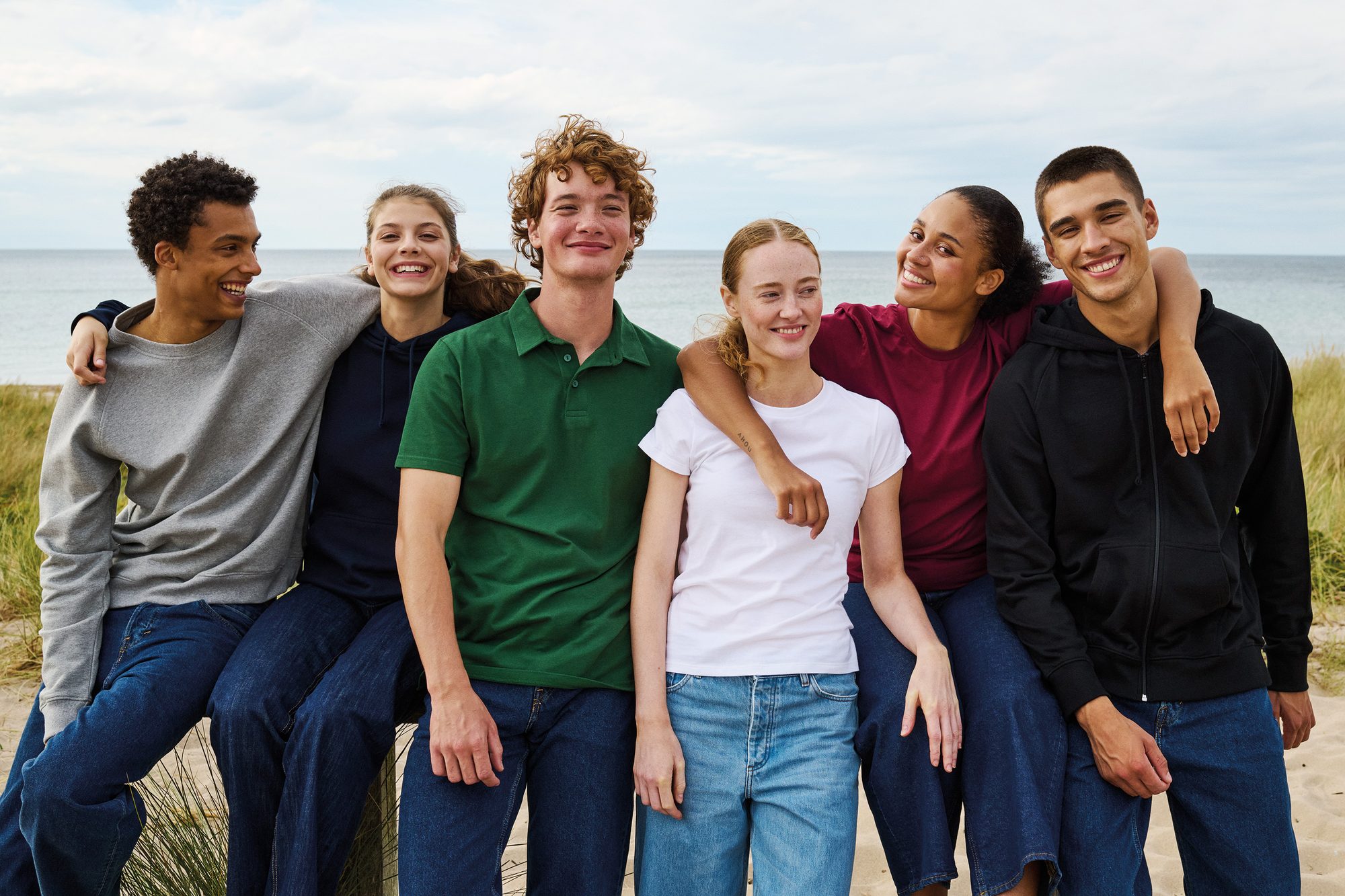 Sechs junge Menschen in PROLINQ Corporate Fashion am Strand · Polos, Hoodies, T-Shirts in sechs Farben